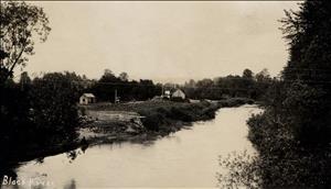 A small river passes farm buildings and a small field with crops growing. Someone sits by the shore in a clearing on the riverbank.